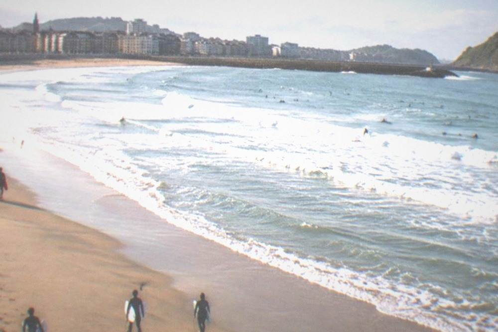 A wide sandy beach with rolling waves and surfers in the water beside a city skyline