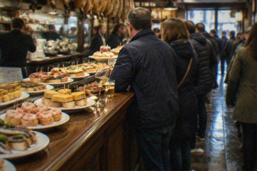 Crowds standing at a pintxo bar counter with small skewered snacks and glasses of wine