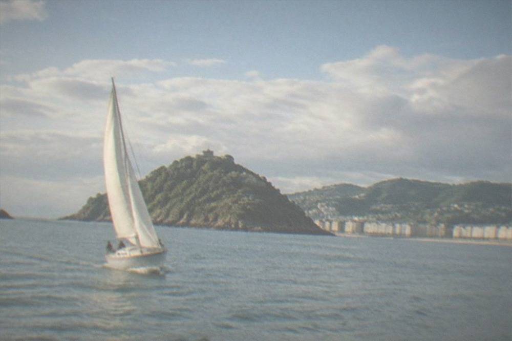 A sailboat heeling gently on the water of La Concha bay with green hills behind