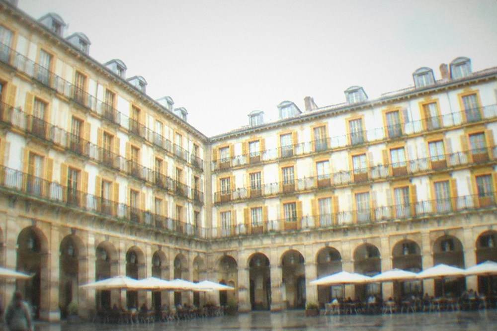 An arcaded plaza with numbered balconies and cafe tables under a cloudy sky