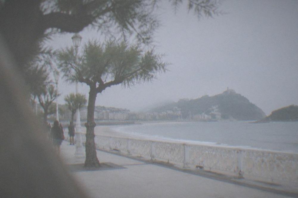 A wide curved bay promenade with a railing, an island offshore and green hills at each end